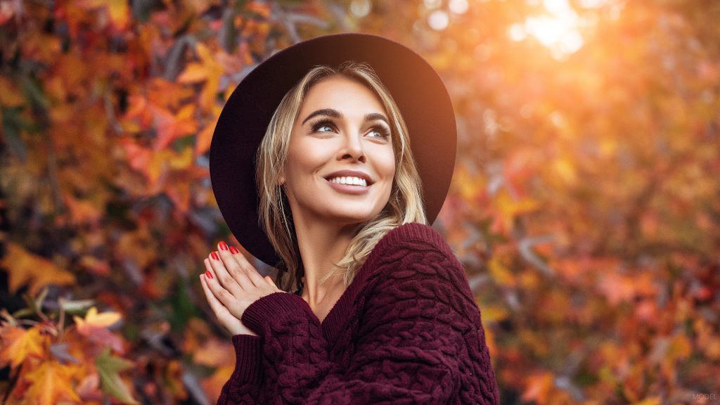 A woman in a hat and sweater posing in front of autumn leaves. (Model)