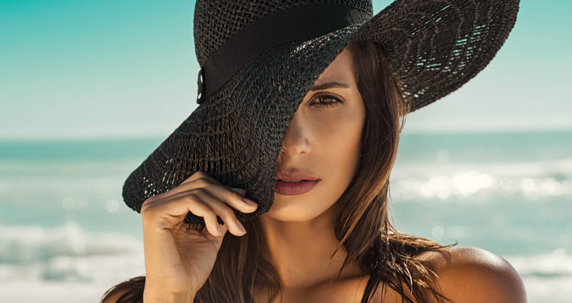 Portrait of fashion young woman posing against sea wearing a black straw hat. Sensual girl covering half face with hat on beach with copy space. Attractive girl looking at camera with sea in background.